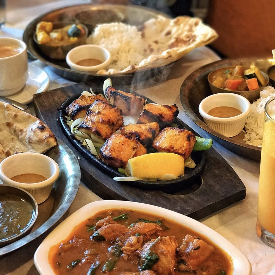 A table laden with various Nepalese dishes, including steaming grilled chicken, curries, rice, and naan.