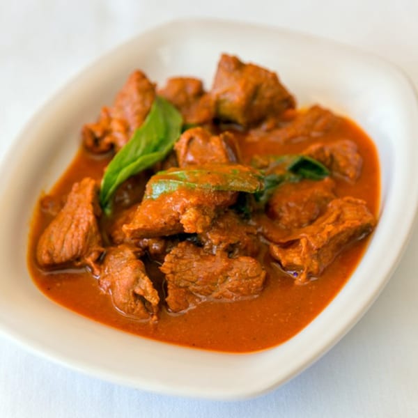 Nepalese lamb curry, a rich red stew with tender meat pieces and green herbs, served in a white bowl.