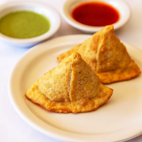 Two golden-brown Nepalese samosas on a white plate, with green and red dipping sauces in bowls.