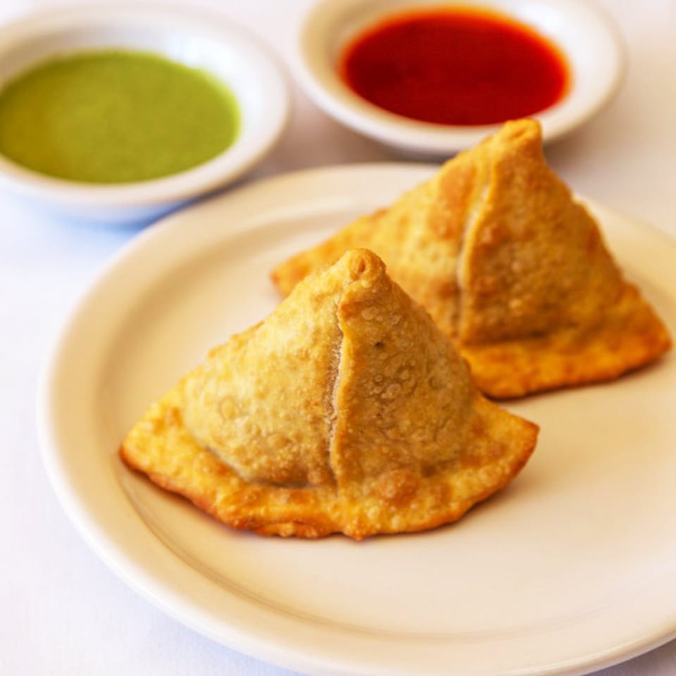 Two golden-brown Nepalese samosas on a white plate, with green and red dipping sauces in bowls.